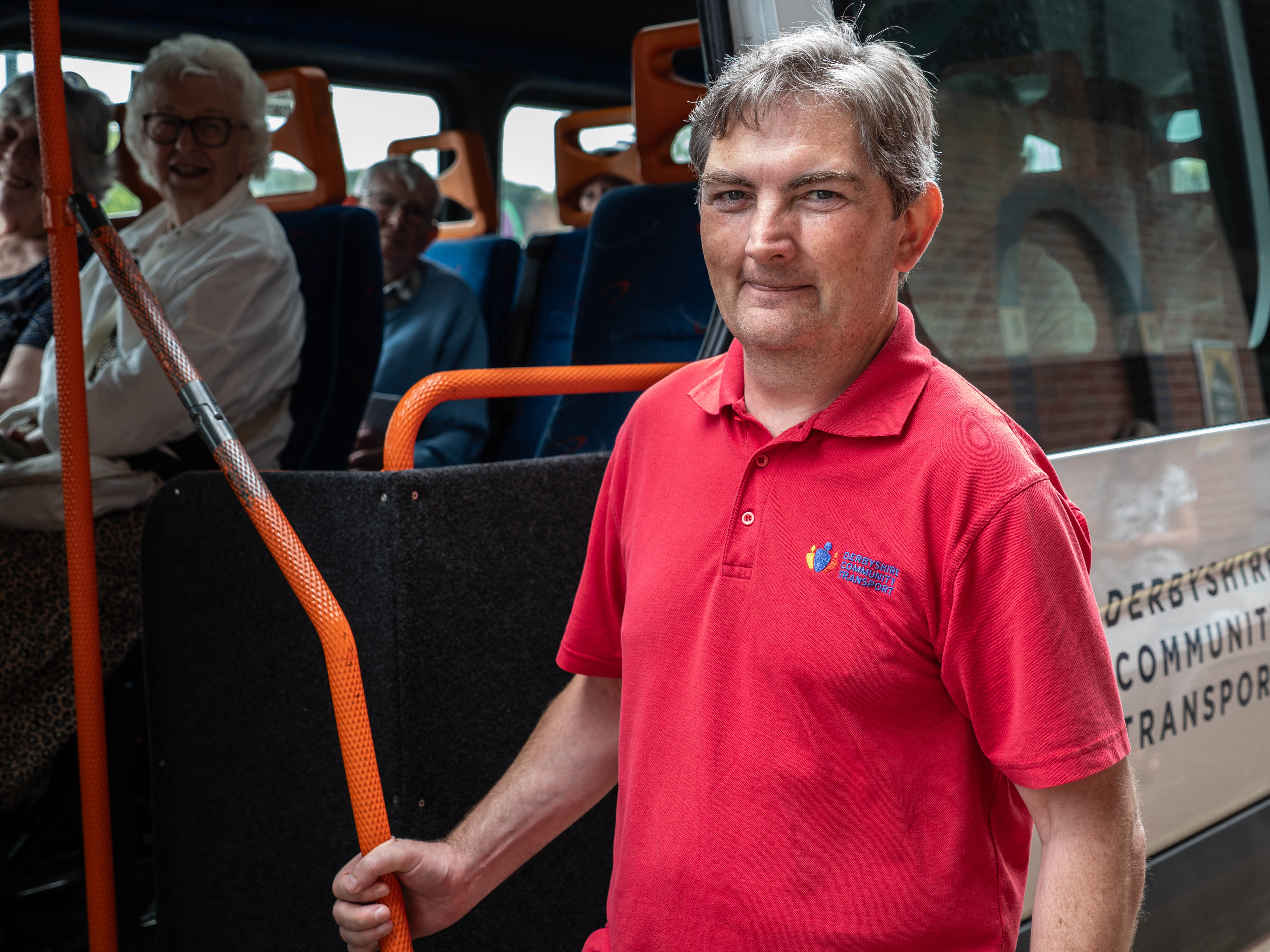 driver in red polo shirt stood in front of an open door of a minibus full of older passengers 