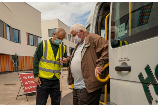 photo of a driver in a high viz vest and face mask assisting an older gentleman getting off a minibus outside a hospital building 