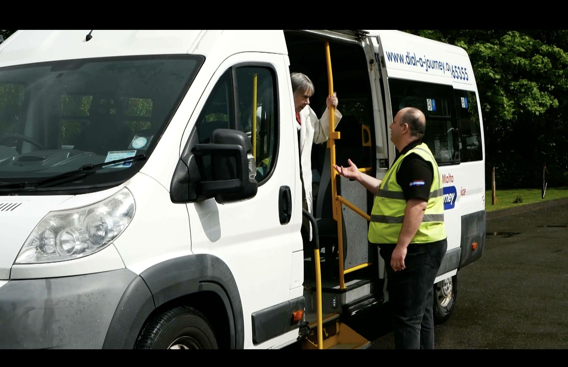 photo of a driver, in high viz jacket, supporting passengers boarding a white minibus.