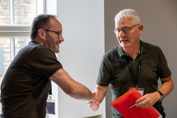 two men in dark coloured t shirts shaking hands. One man is holding a red folder. 