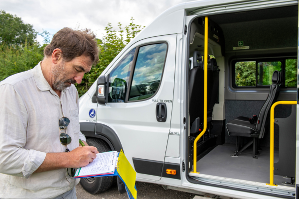 man in white shirt writing on paper stood next to a minibus.