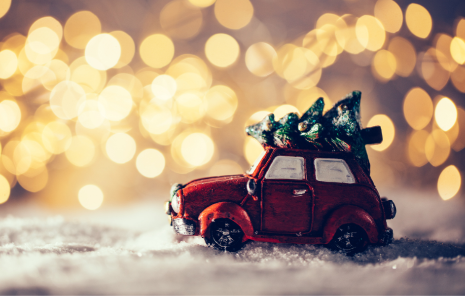A small red toy car carrying a Christmas tree on its roof sits on a snowy surface, with warm, out-of-focus festive lights glowing in the background.