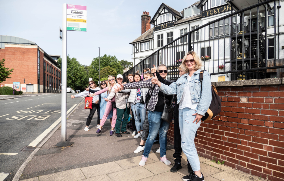 A group of women stand in a line at a roadside bus stop, smiling and giving thumbs up toward the camera, with a traditional black-and-white pub building and urban street in the background.