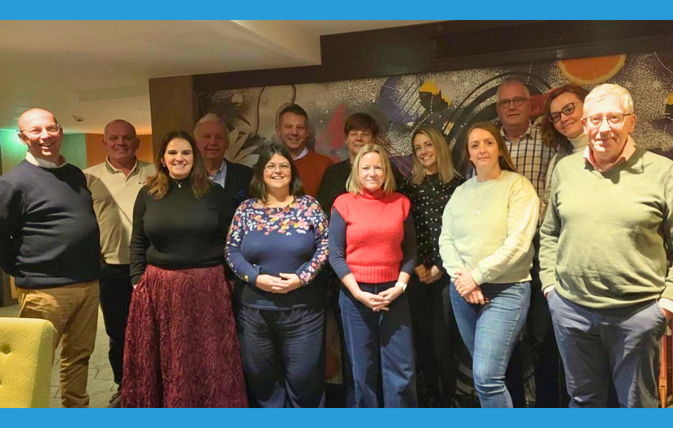 A group of around 14 people standing side by side in a meeting room, smiling at the camera. They are casually dressed and positioned in front of a colourful abstract mura