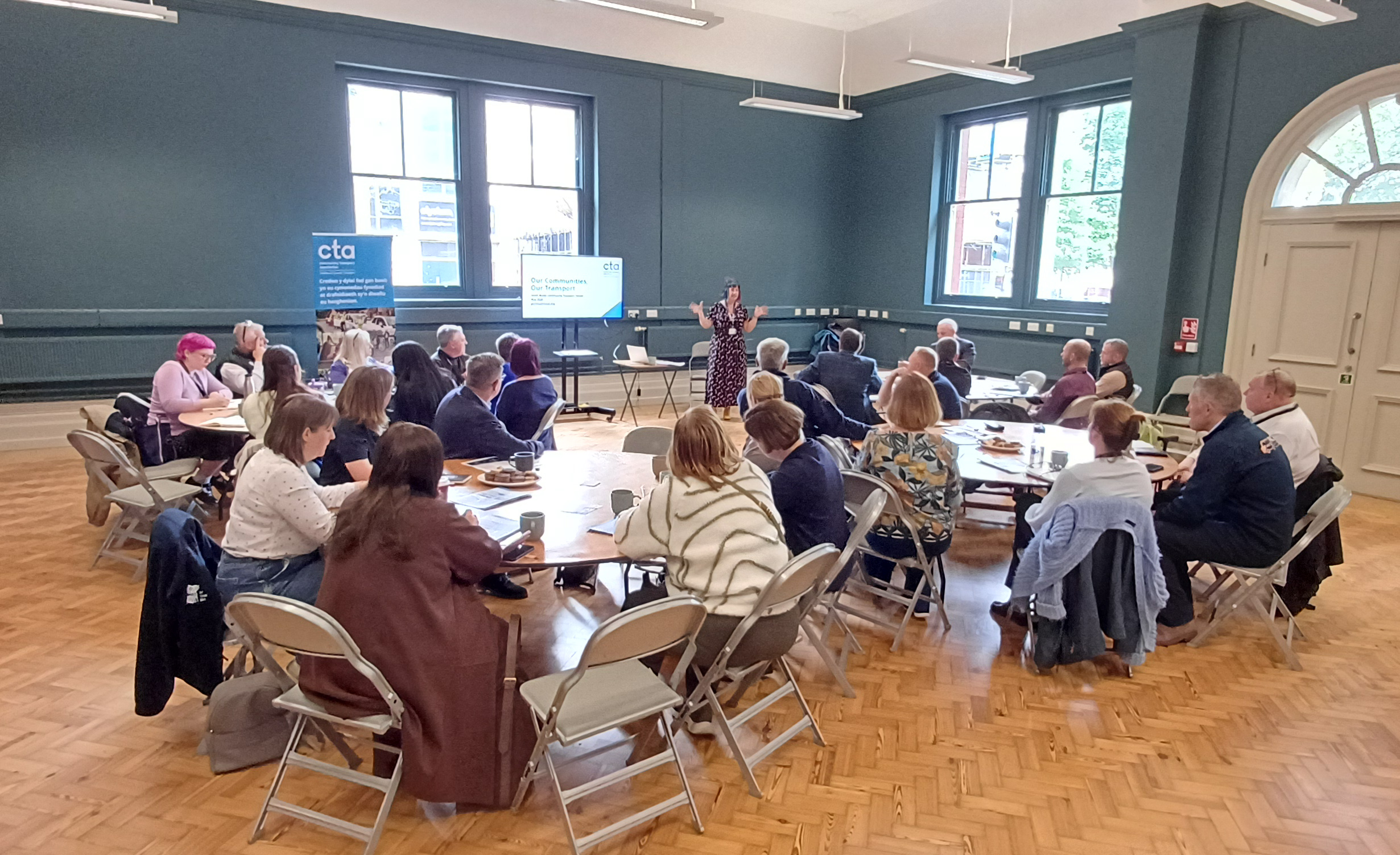 Image of various people in a room with wooden flooring, sitting at circular tables on folding chairs watching a presentation and speaker