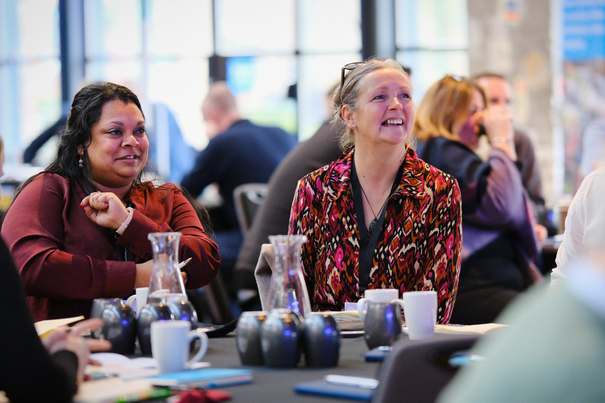 photo shows two women sat at a conference table watching a speaker present. 