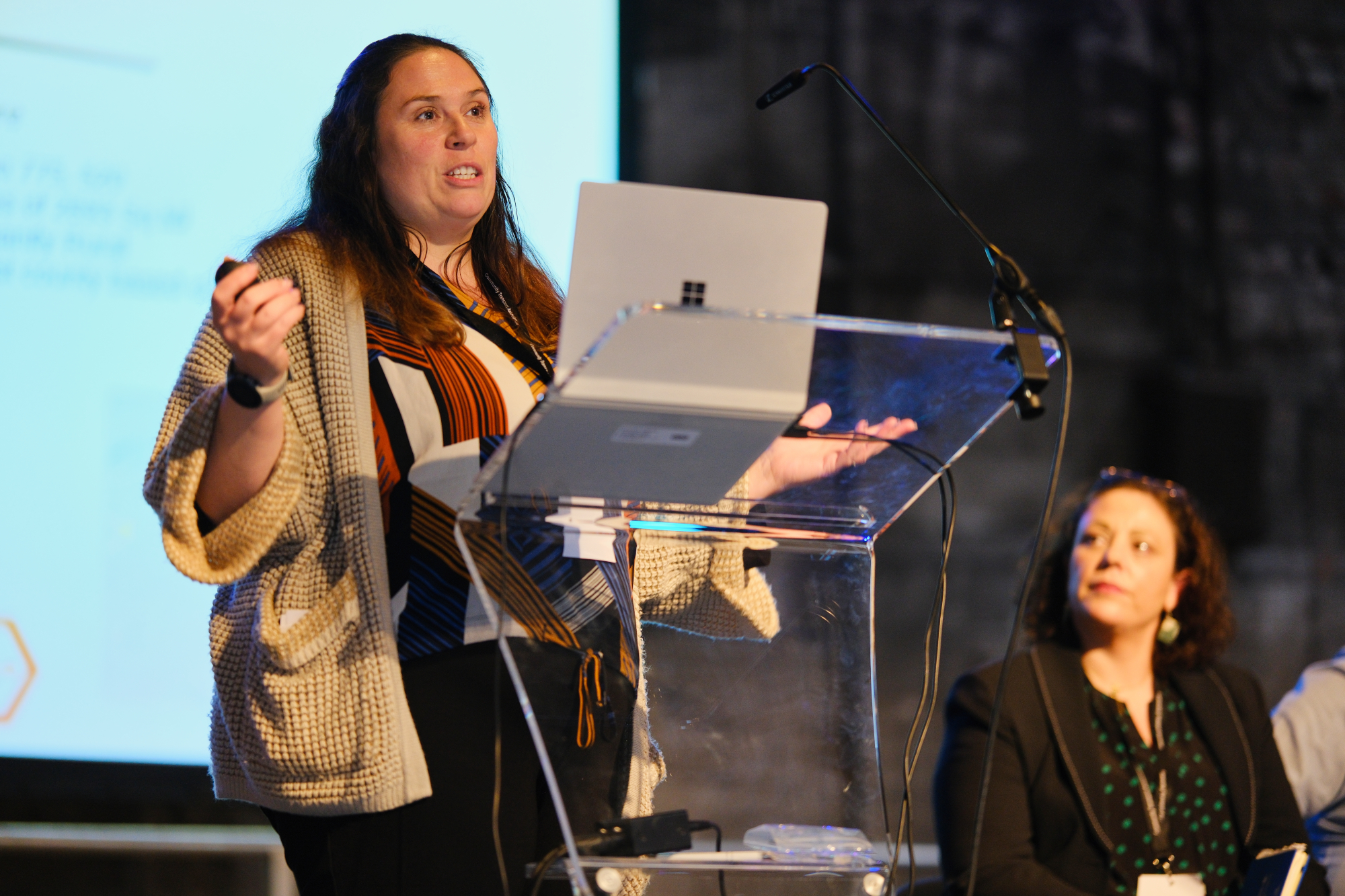 photo shows a woman in a cream woven cardigan and black trousers talking at a lectern with a woman in black sat behind watching 