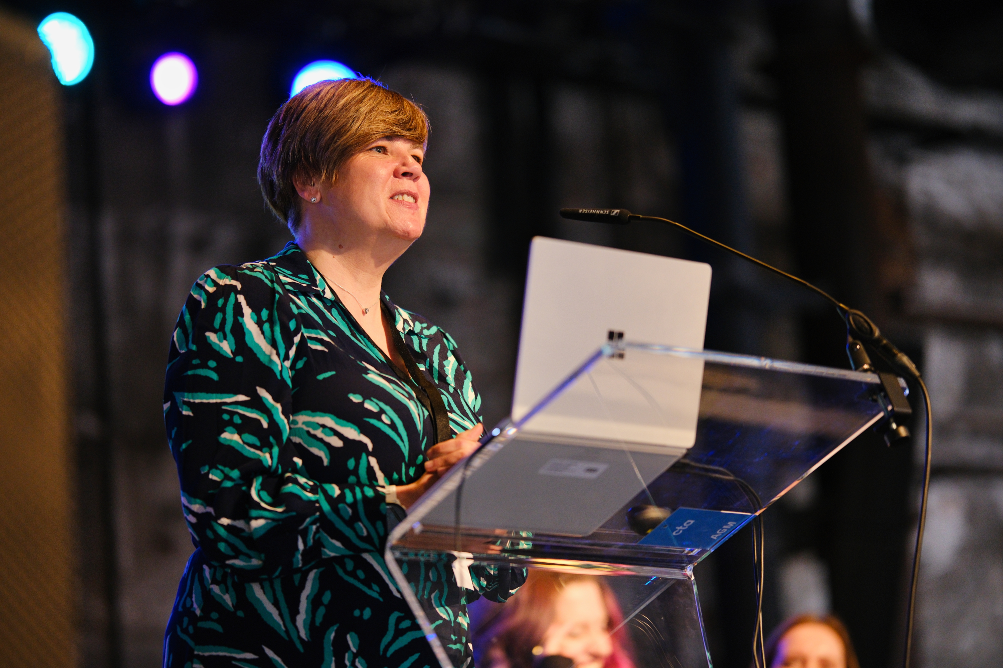 photo shows a woman in a navy, green and white dress, with short cropped brown hair talking at a glass lectern.