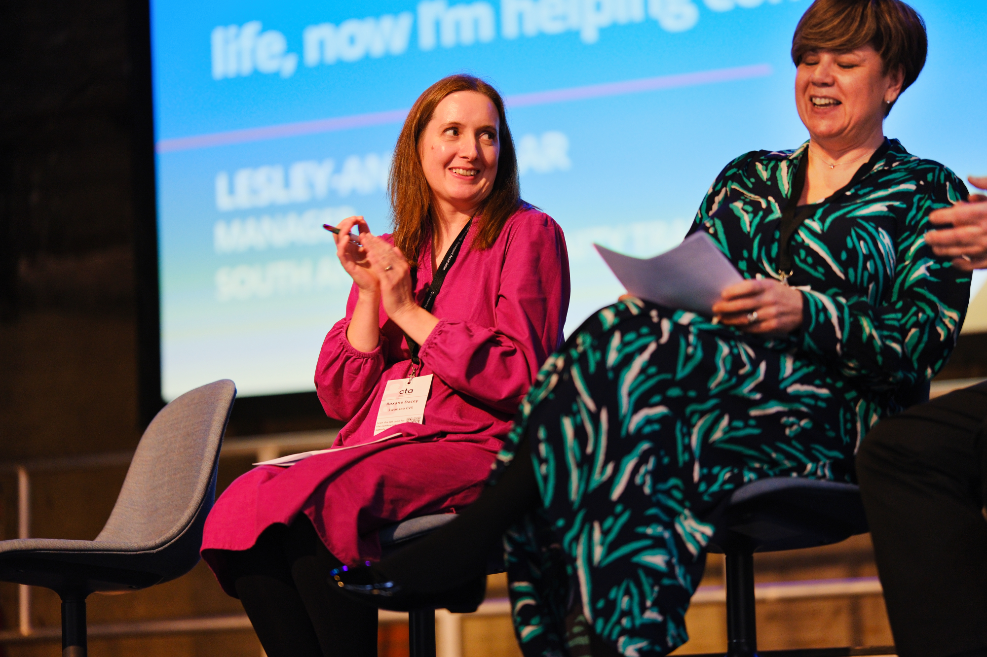 photo shows a woman in a navy, green and white dress, with short cropped brown hair laughing, while a woman in a pink dress, sitting next to her, claps.