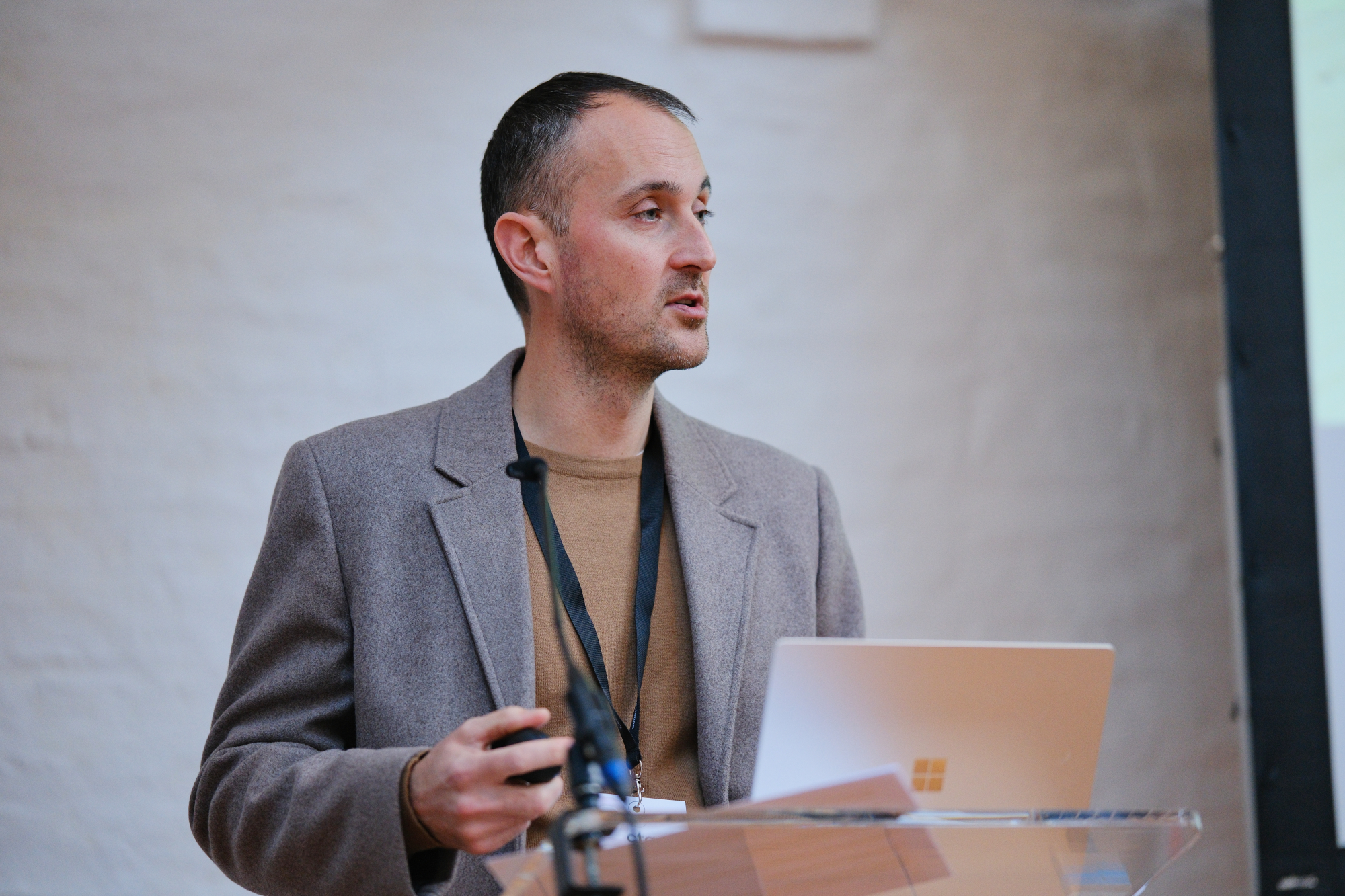 photo shows a man with dark hair, grey coat and lanyard presenting at a glass lectern