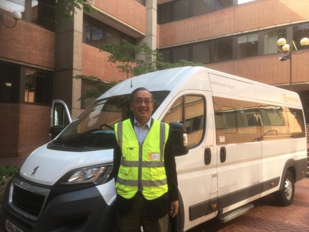 An image of Tim Sai Louie in a hi-vis vest standing outside a white community transport vehicle