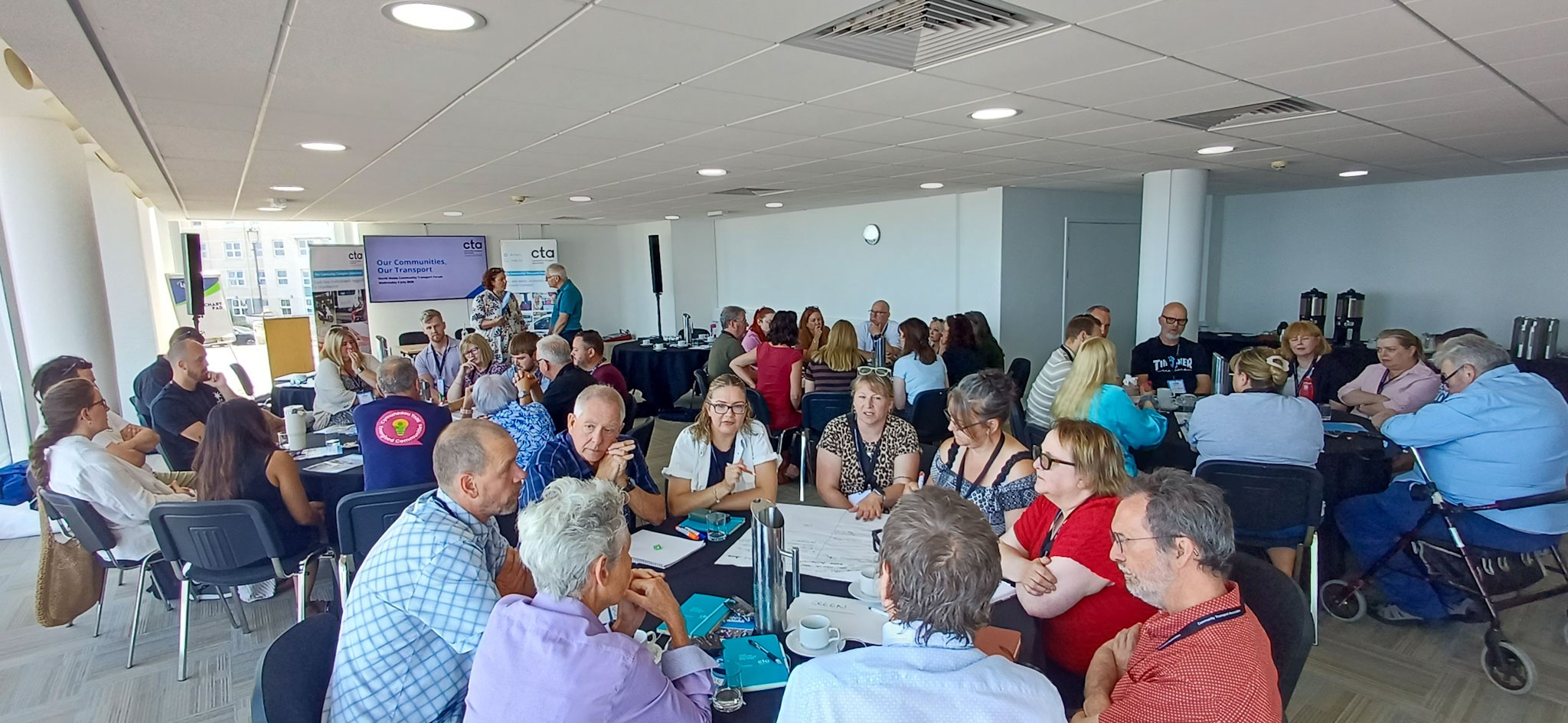 An image of a room filled with participants with a presentations screen in the background and CTA branded banners.