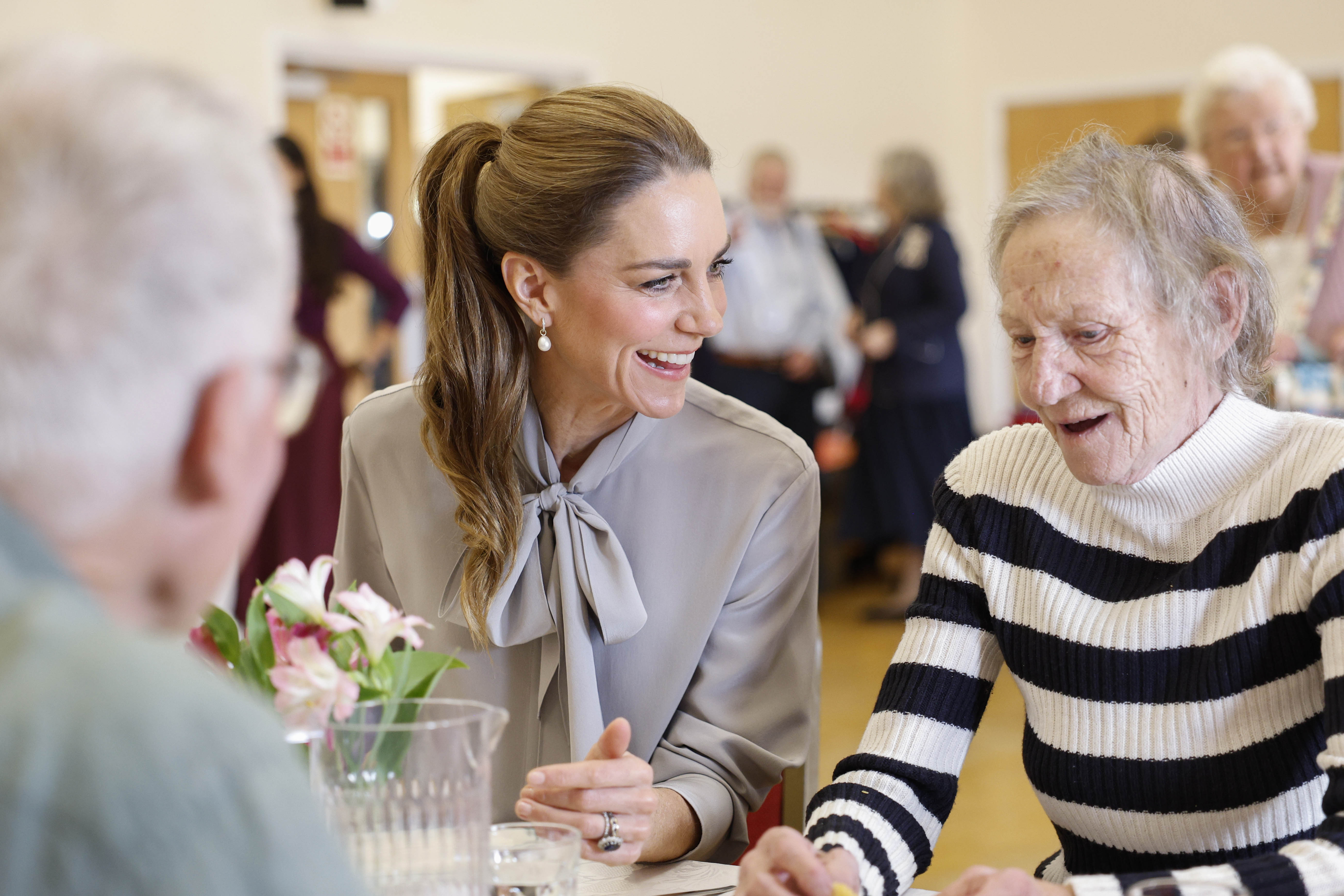 The Princess of Wales speaking to an older person while sitting down in a hall attending a lunch club
