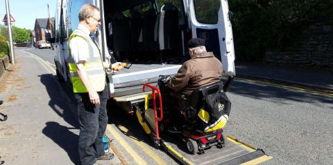 photo showing a driver in hi viz jacket assisting a man in a wheelchair to access a minibus.