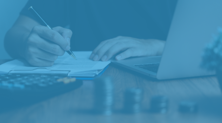 Person writing on a clipboard with a pen, next to a laptop and stacked coins in the foreground, symbolizing financial planning and funding