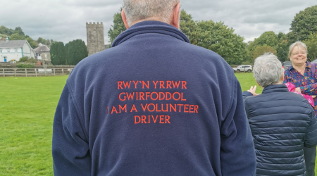  A person wearing a navy jacket with red text on the back that reads "RWY'N YR WR GWIRFODDOL I AM A VOLUNTEER DRIVER" in both Welsh and English. They are standing outdoors in a grassy area, with a stone tower and trees visible in the background. Two other people are also present, one facing away from the camera and another smiling towards it.