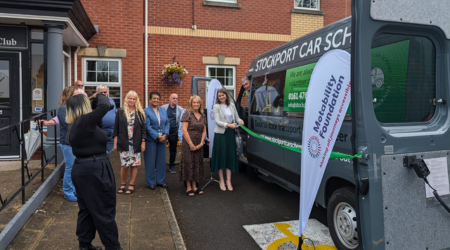 Group of people standing in front of a van with 'Stockport Car Scheme' written on it. The group includes men and women, and they are posing for a photo while a photographer captures the moment. Two 'Motability Foundation' banners are placed next to the van outside a brick building.