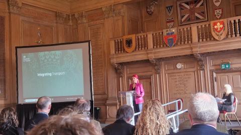 photo shows Transport Secretary, Louise Haig, dressed in pink, addressing a room of people, in front of a large screen. 