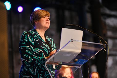 photo shows a woman in a navy, green and white dress, with short cropped brown hair, speaking at a glass lectern