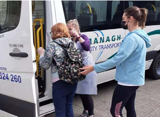 Photo shows two women supporting another woman boarding a white mini bus. 