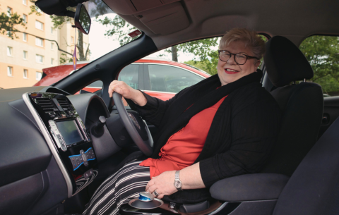 A smiling woman sits in the driver’s seat of a parked car, wearing glasses, a red top and a black cardigan. She has one hand on the steering wheel and looks relaxed and confident, ready to drive.