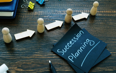 Wooden figures and arrow cut-outs arranged in a line on a dark wooden desk, symbolising progression or succession. Nearby are black cards with “Succession Planning” written in white marker, alongside a pen and office supplies.