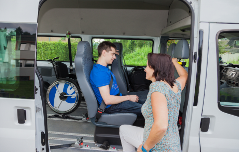 A community transport vehicle with its side door open. A passenger is seated and secured inside, and a support worker kneels beside him, chatting and ensuring he is comfortable. A wheelchair is secured behind them.