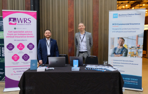 Two representatives stand behind an information table at an indoor event, flanked by branded banners for WRS Insurance Brokers and Business Choice Direct Insurance Services. The table displays laptops, brochures, and pens, with the banners highlighting specialist insurance services for charities, social enterprises, and commercial organisations.