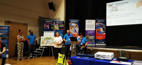 Image of speakers standing at the front of the room in a large hall with PACTO banners and other informative material.
