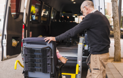 An image of a community transport vehicle opened at the back with a ramp, where an individual is helping another individual in a wheelchair enter the vehicle using the ramp