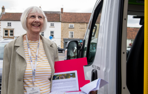 Image of a smiling person with a clipboard, wearing a volunteer lanyard and standing outside of a community transport vehicle