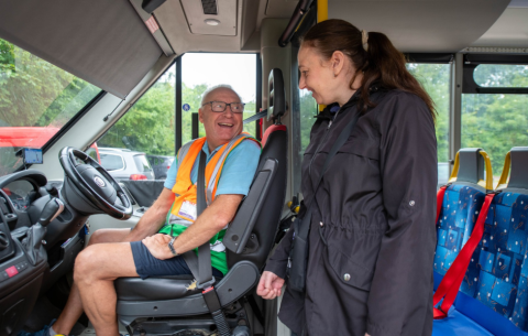 Image of two people in a community transport vehicle, with the driver wearing a hi vis vest looking at another person who appears to be smiling at him