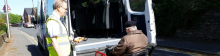 photo showing a driver in hi viz jacket assisting a man in a wheelchair to access a minibus.