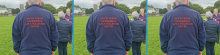  A person wearing a navy jacket with red text on the back that reads "RWY'N YR WR GWIRFODDOL I AM A VOLUNTEER DRIVER" in both Welsh and English. They are standing outdoors in a grassy area, with a stone tower and trees visible in the background. Two other people are also present, one facing away from the camera and another smiling towards it.